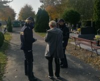 na alejce cmentarza dwie umundurowane policjantki rozmawiają z kobietą, która jest widoczna od tyłu, po obu stronach alejki znajdują się nagrobki