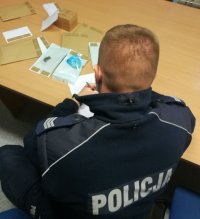 W pomieszczeniu służbowym umundurowany policjant wykonuje oględziny zabezpieczonych narkotyków, na stole leżą zapakowane koperty z narkotyki