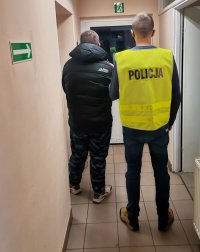 na korytarzu komisariatu policjant ubrany w kamizelkę odblaskową z napisem POLICJA prowadzi zatrzymanego który ma założone kajdanki na ręce trzymane, z prawej strony widać drzwi