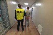 korytarz komendy, nieumundurowany policjant w żółtej kamizelce z napisem policja prowadzi zatrzymanego mężczyznę