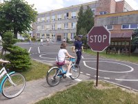 Przed budynkiem szkoły dziecko na rowerze pokonuje rowerowy tor przeszkód, obok stoi umundurowany policjant