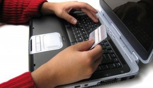 Na obrazku widoczne są dłonie osoby, która znajduje się przed laptopem. W jednej ręce trzyma kartę płatniczą  a drugą pisze na klawiaturze urządzenia.