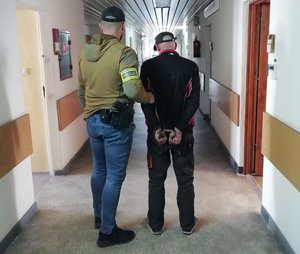 Na korytarzu budynku komendy nieumundurowany policjant z opaską z napisem Policja prowadzi zatrzymanego który ma założone kajdanki.