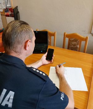Policjant siedzący przy biurku trzyma w lewej dłoni telefon komórkowy, w prawej długopis sporządzając dokumentację
