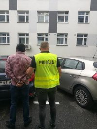 przed budynkiem komendy policjant prowadzi zatrzymanego do nieoznakowanego radiowozu, zatrzymany ma założone kajdanki na ręce trzymane z tyłu