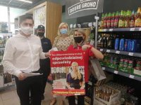 wizyta w sklepie,członek komisji oraz sprzedawca trzymają rozwinięty plakat, z tyłu stoi policjant i drugi sprzedawca