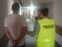 POLICJANTKA UBRANA W KAMIZELKĘ ODBLASKOWĄ Z NAPISEM POLICJA PROWADZI ZATRZYMANEGO KTÓRY MA ZAŁOŻONE KAJDANKI NA RĘCE TRZYMANE  Z TYŁU