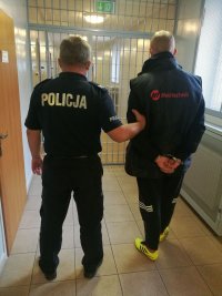 na korytarzu w budynku komendy umundurowany policjant prowadzi zatrzymanego, który ma złożone kajdanki na ręce trzymane z tyłu, przed  nimi widoczna jest metalowa krata, po lewej stronie drzwi od pomieszczenia