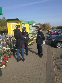 na chodniku przez cmentarzem przed stoiskiem z zniczami i kwiatami  dwie umundurowane policjantki, które mają osłonięte twarze maseczkami, rozmawiają z kobietą, która stoi tyłem, obok parking, gdzie stoją zaparkowane samochody, obok blaszanej budki stoją kwiaty i znicze