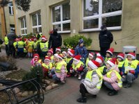 przed budynkiem Przedszkola nr 15 w Sieradzu grupa kilkudziesięciu dzieci wspólnie z policjantami udekorowała drzewka elementami odblaskowymi. Policjanci stoją przy udekorowanych drzewkach,  ubrani są w granatowe mundury na głowach maja czapki Mikołaja, dzieci kucają przed drzewkami mają założone kamizelki odblaskowe, na głowach czapki Mikołaja,