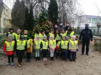 na placu  Przedszkola nr 4 w Sieradzu grupa przedszkolaków wspólnie z policjantami i pracownikiem WORD  stoi przed udekorowana choinka elementami odblaskowymi. Policjanci ubrani są w granatowe mundury na głowach maja czapki Mikołaja, dzieci mają założone kamizelki odblaskowe, z tyłu w oddali widać bloki mieszkalne