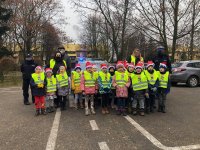 przed budynkiem Przedszkola nr 15 w Sieradzu grupa kilkudziesięciu dzieci wspólnie z policjantami stoją przed oznakowanym radiowozem z włączonymi sygnałami swietlnymi  policjanci ubrani są w granatowe mundury na głowach mają czapki Mikołaja, dzieci  mają założone kamizelki odblaskowe, na głowach czapki Mikołaja,