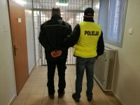 na korytarzu policyjnego aresztu policjant ubrany w kamizelkę odblaskową z napisem policja prowadzi zatrzymanego, kory ma założone kajdanki na ręce trzymane z tyłu, z lewej strony widać drzwi do pomieszczenia , z przodu kratę, po prawej stronie zakratowane okno