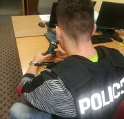 w pomieszczeniu służbowym przy biurku siedzi odwrócony tyłem policjant, ubrany  kamizelkę z napisem POLICJA trzyma w dłoniach odzyskany z kradzieży laser krzyżowy, na biurku widac dwa monitory