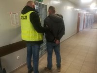 w areszcie policjant ubrany w kamizelkę odblaskowa z napisem POLICJA prowadzi zatrzymanego który ma założone kajdanki na ręce trzymane z tyłu, z przodu widać  kratę na drzwiach