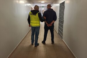 zdjęcie przedstawia korytarz policyjnego aresztu. na pierwszym planie widzimy nieumundurowanego policjanta, w żółtej kamizelce odblaskowej z napisem Policja, który prowadzi zatrzymanego mężczyznę. Zatrzymany ma założone kajdanki.