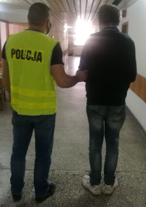 na korytarzu w budynku komendy policjant ubrany w kamizelkę odblaskową z napisem policja prowadzi zatrzymanego, który ma założone kajdanki na ręce trzymane z przodu z prawej strony widać drzwi do pomieszczenia ,