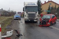 na drodze asfaltowej stoi ciężarowa biała  scania, z prawej strony czerwony samochód osobowy z uszkodzonym przodem, z lewej strony na poboczu oznakowany radiowóz, za nim oznakowany wóz straży pożarnej, widać kilku umundurowanych strażaków, z przodu pozrzucane elementy zderzaka