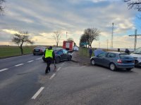 Zdjęcie przedstawia miejsce  wypadku drogowego.Jest to skrzyżowanie dróg na którym doszło do zderzenia się dwóch pojazdów. Na pierwszym planie widać policjanta w ubraniu cywilnym, który ma na sobie kamizelkę odblaskową z napisem ,,Policja&quot;.Który wykonuje czynności na miejscu zdarzenia. Tuż przed nim widoczne są dwa pojazdy, które brały  udział w zdarzeniu i posiadają uszkodzenia karoserii w przednich częściach pojazdu, oraz dwa  pojazdy, które stoją na drodze oraz na poboczu, a przy nich stojące osoby. Na drugim planie w oddali widać oznakowane pojazdy straży pożarnej oraz policji stojące na poboczu.