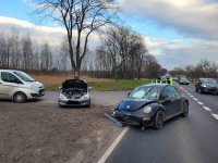 Zdjęcie przedstawia miejsce zdarzenia drogowego z udziałem dwóch pojazdów osobowych na skrzyżowaniu dróg. Na pierwszym planie widoczne są dwa pojazdy osobowe z widocznymi uszkodzeniami karoserii w przednich ich częściach. Na drugim planie w oddali widoczne są pojazdy  przejeżdżające obok miejsca zdarzenia. Tuż za jednym z uszkodzonych pojazdów widoczne są dwie osoby, jedna z nich to umundurowany funkcjonariusz Wydziału Ruchu Drogowego, który na głowie ma białą czapkę, a drugi to nieumundurowany funkcjonariusz policji, który posiada na sobie kamizelkę odblaskową.