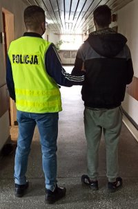 korytarz komendy, policjant w żółtej kamizelce odblaskowej prowadzi zatrzymanego mężczyznę