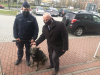 Przed budynkiem Urzędu Gminy Sieradz stoi umundurowany policjant trzymający psa na smyczy. Obok nich pochylony nad psem stoi Wójt Giny Sieradz. Mężczyźni mają założone maseczki na twarzy. W tle widoczny jest parking, na którym stoją samochody.