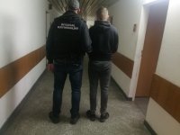 na korytarzu w budynku komendy nieumundurowany policjant, który ma założoną czarną kamizelkę taktyczną z napisem POLICJA prowadzi zatrzymanego mężczyznę, obydwaj są odwróceni tyłem