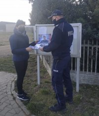 Przed tablicą ogłoszeniową znajduje się umundurowany funkcjonariusz Policji, wręczający materiały promocyjne sołtysowi.