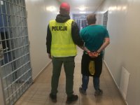w areszcie policjant ubrany w kamizelkę odblaskowa z napisem POLICJA prowadzi zatrzymanego który ma założone kajdanki na ręce trzymane z tyłu, z lewej strony widać kratę