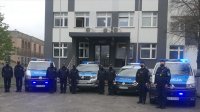 przed budynkiem komendy stoją radiowozy oznakowane z włączonymi śwatłami obok umundurowani policjanci