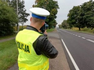 na obrazku widoczny jest stojący przy jezdni policjant, który dokonuje pomiaru prędkości nadjeżdżającego pojazdu