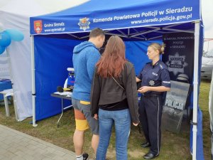 Przy namiocie  z logo Komendy Powiatowej Policji w Sieradzu  stoi umundurowana policjantka która rozmawia z kobietą i mężczyzną,   na stoliku w namiocie leżą ulotki profilaktyczne za stołem banery Komendy Powiatowej Policji w Sieradzu