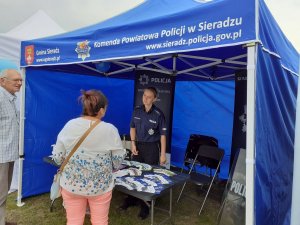 Przy namiocie  z logo Komendy Powiatowej Policji w Sieradzu  stoi umundurowana policjantka która rozmawia z kobietą i mężczyzną,   na stoliku w namiocie leżą ulotki profilaktyczne za stołem banery Komendy Powiatowej Policji w Sieradzu