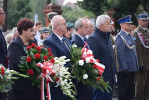 DELEGACJA SŁUŻB MUNDUROWYCH NA UROCZYSTOŚCI, UMUNDUROWANY STRAŻAK, POLICJANT, STRANIK MIEJSKI, DWIE OSOBY TRZYMAJĄ KWIATY