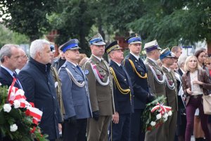 DELEGACJA SŁUŻB MUNDUROWYCH NA UROCZYSTOŚCI, UMUNDUROWANY STRAŻAK TRZYMA KWIATY, OBOK NIEGO UMUNDUROWANY POLICJANT, STRAŻNIK MIEJSKI