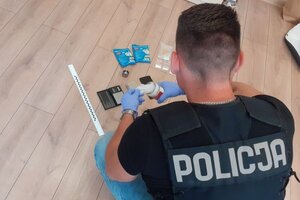 W pomieszczeniu służbowym na podłodze  policjant ubrany w czarną kamizelkę z napisem policja widziany od tyłu pochyla się nad rozłożonymi środkami odurzającymi, widoczne są torebki z białym proszkiem, waga, niebieskie  opakowania po produktach spożywczych