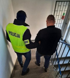 Policjant który ma założoną kamizelkę odblaskową prowadzi prowadzi zatrzymanego