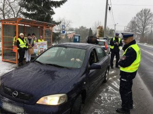 Dzieci wspólnie z policjantami przeprowadzają kontrole drogowe kierowców i wręczają woreczki  z ziemią  lub uśmiechnięte zawieszki, dzieci trzymają w rękach plakat akcji