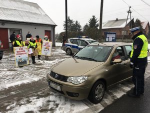 Dzieci wspólnie z policjantami przeprowadzają kontrole drogowe kierowców i wręczają woreczki  z ziemią  lub uśmiechnięte zawieszki, dzieci trzymają w rękach plakat akcji