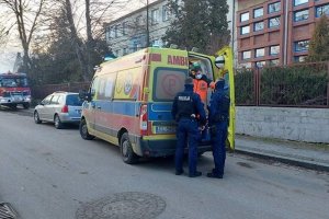 na zdjęciu widoczna jest zaparkowana na jezdni karetka pogotowia; tylne drzwi karetki są otwarte, stoi przy nich dwóch umundurowanych policjantów; w tle znajduje się budynek szkoły