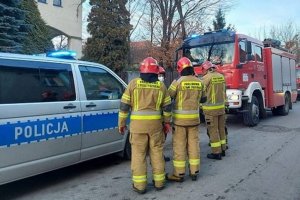 na zdjęciu widoczne są zaparkowane na ulicy pojazdy służb ratowniczych tj. straży pożarnej i policji; na ulicy stoją również strażacy