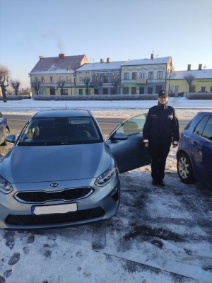 na zdjęciu widoczna jest umundurowana funkcjonariuszka Policji stojąca obok samochodu; w tle znajdują się kamienice Rynku w Warcie