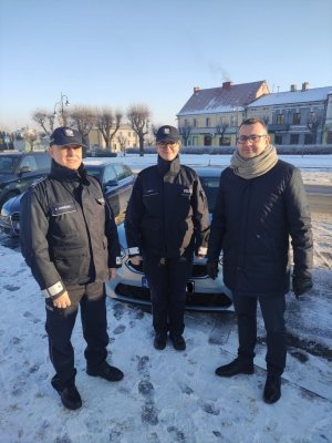 na zdjęciu widoczne są trzy osoby, w tym dwóch umundurowanych funkcjonariuszy policji stojących przed samochodem; w tle zabudowania rynku w Warcie