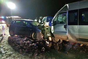 na zdjęciu widoczne są dwa pojazdu - osobowy i bus; pojazdy stoją na jezdni, przodem do siebie, są uszkodzone po zderzeniu; jest noc; w tle znajduje się kilka postaci, w tym strażacy