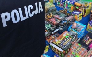 na obrazku znajduje się fragment postaci umundurowanego policjanta z widocznym na kurtce napisem &quot;POLICJA&quot;, na drugim planie widać stoisko z fajerwerkami