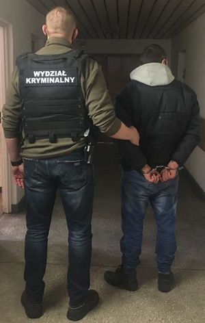 na korytarzu budynku komendy policjant ubrany w czarną kamizelkę z napisem Wydział Kryminalny prowadzi zatrzymanego który ma założone kajdanki na ręce trzymane z tyłu