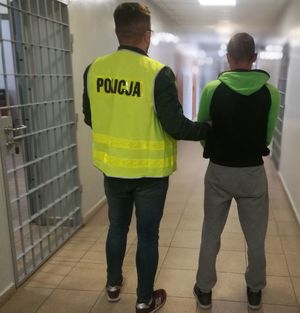 w areszcie policjant ubrany w kamizelkę odblaskowa z napisem POLICJA prowadzi zatrzymanego który ma założone kajdanki na ręce trzymane z przodu, z prawej strony widać kratę na oknie, z lewej drzwi do pomieszczeń