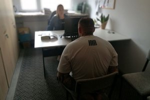 w pokoju służbowym policjantka przesłuchuje zatrzymanego który siedzi tyłem , na rękach ma założone kajdanki
