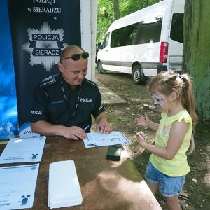 podczas pikniku przy namiocie policyjnym przy stoliku  umundurowany policjant wykonuje odciski palców