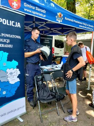 odczas pikniku przy namiocie policyjnym chłopiec przymierza elementy wyposażenia do służby obok stoi umundurowany policjant,